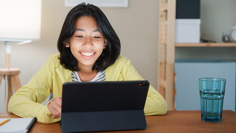 A teenage girl sits at a desk, looking at an tablet screen and smiling