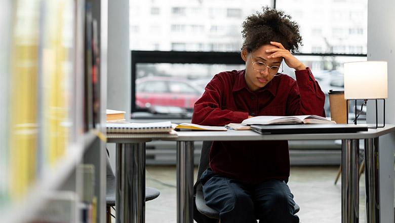 A teenager studies a book at a desk with her head in her hand