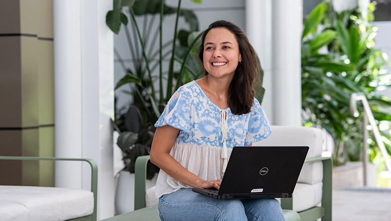 Barbara Azevedo de Oliveira sits with a laptop smiling out the front of UQ's Student Central, surrounded by green plants and white pillars
