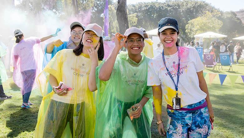 Students in colourful ponchos smile at the camera as coloured powders are thrown in the air around them