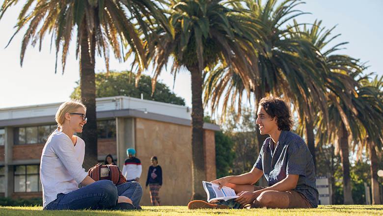 Two students sit on green grass chatting to one another in the sunshine in UQ Gatton campus' Central Walkway