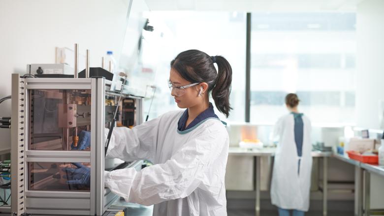 A UQ Researcher in a lab coat and safety glasses handles equipment in a lab