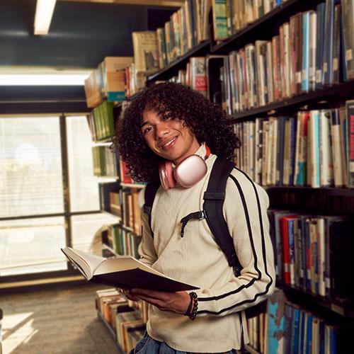 A non-binary student stands in a library with a book