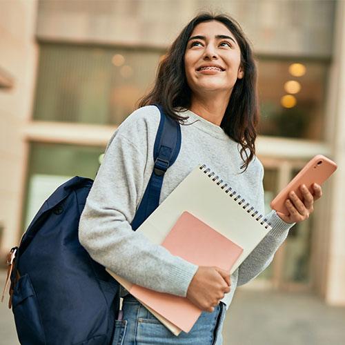 A female student stands wearing a backpack and holding a notebook and phone