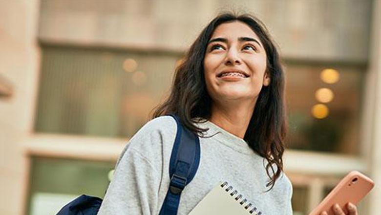 A female student stands wearing a backpack and holding a notebook and phone