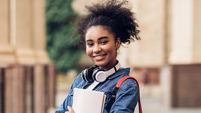 A female student stands in university cloisters wearing a backpack and headphones and holding a notebook