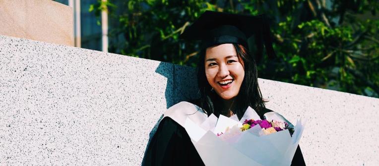 Graduating student wearing a gown and mortarboard and holding a bunch of flowers