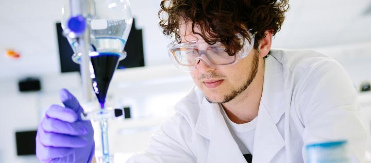 Student working in a lab environment and wearing a lab coat, protective glasses and gloves.