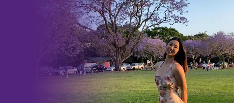 Missy stands in front of a green field, with jacaranda trees in the background
