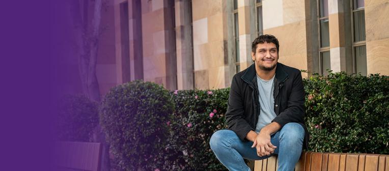 UQ student Jamaine Wilesmith sits in The Great Court with shrubbery and sandstone buildings in the background