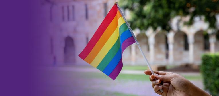 a hand holds a rainbow flag in front of UQ St Lucia's sandstone cloisters