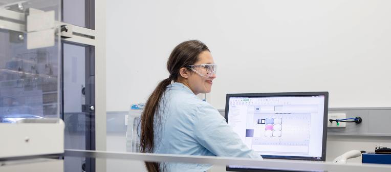 A UQ student in a lab coat and safety glasses sits in front of a computer in a lab