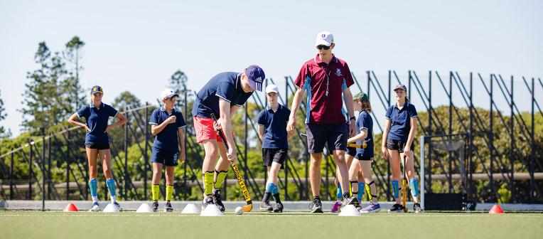 A UQ physical education student teaches a school students how to play hockey