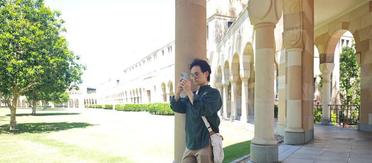 Male student leaning on a cloister taking a photo with his phone