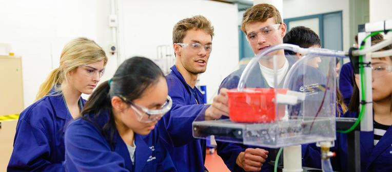 A group of UQ engineering students in lab coats and safety glasses inspect an experiment