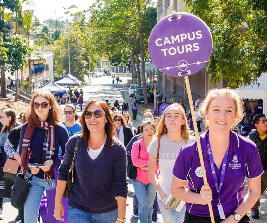 A student in a purple UQ shirt holds a "Campus tours" sign and leads a group of prospective students around St Lucia