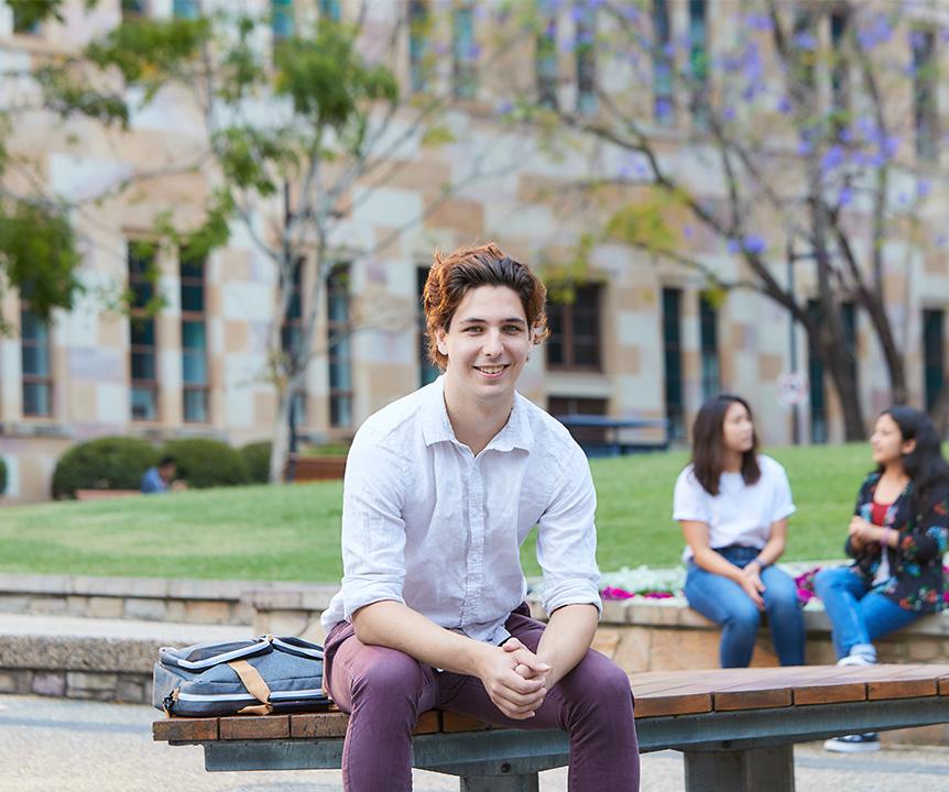 Student sitting on bench with trees and a sandstone building in the background.