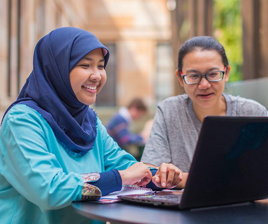 Two women are smiling, sitting at a table looking at a laptop