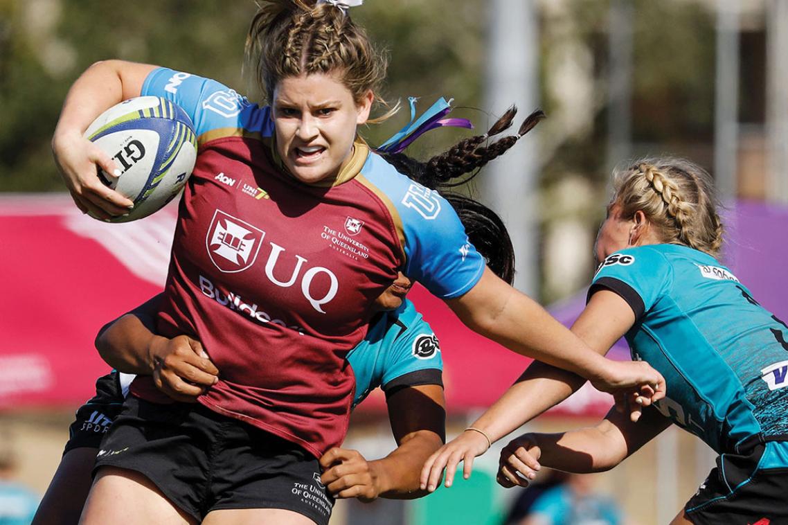 Student in blue and maroon UQ jersey holding the football and dodging a tackle in a game of women's rugby.