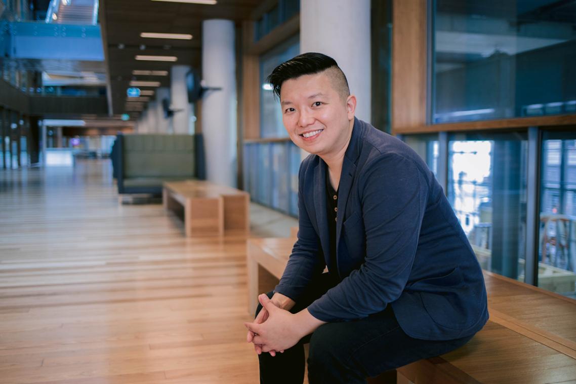 Professor Ryan Ko sits with hands interlinked and smiling on a wooden bench indoors