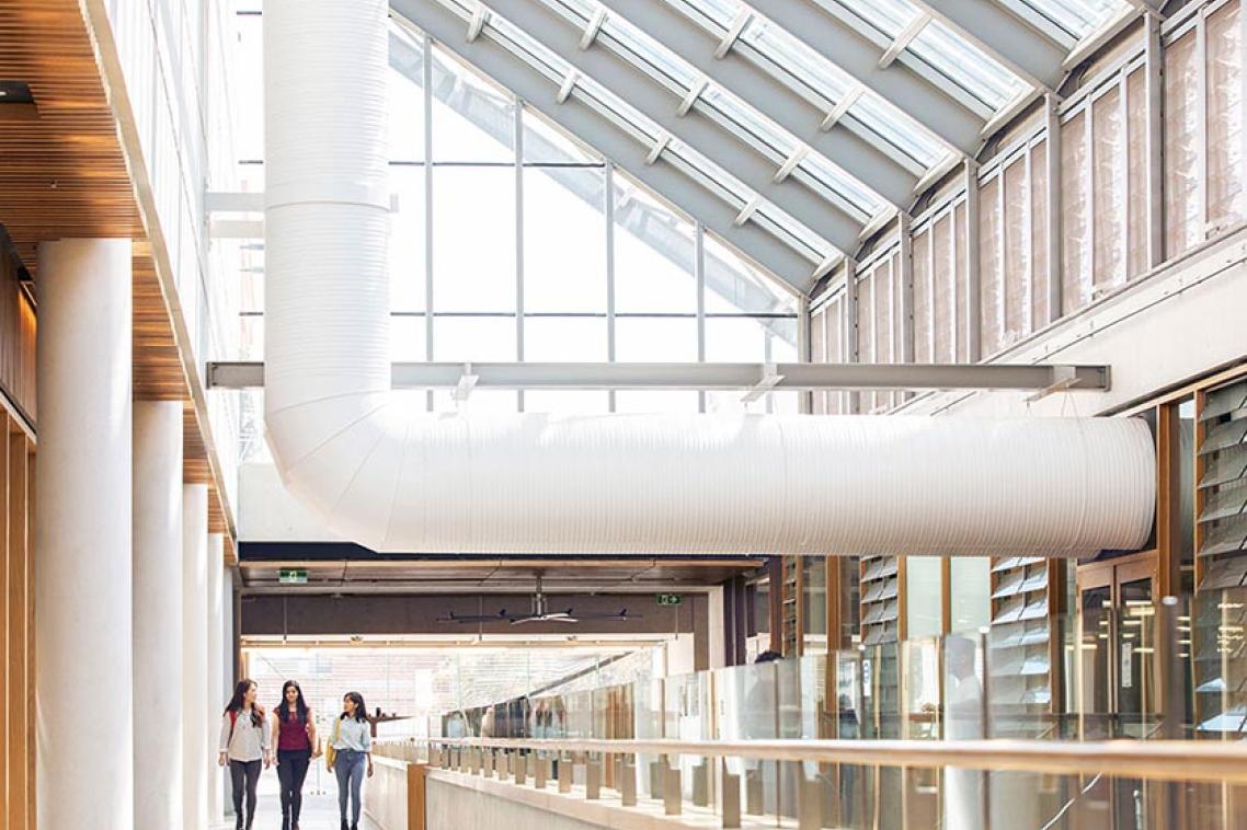 Students walking through the vast interior of the Advanced Engineering Building. Sunlight pours through the glass ceiling and a large industrial pipe runs over their heads.