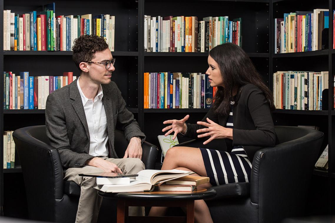 Two students talking at a table with bookshelves behind them. 