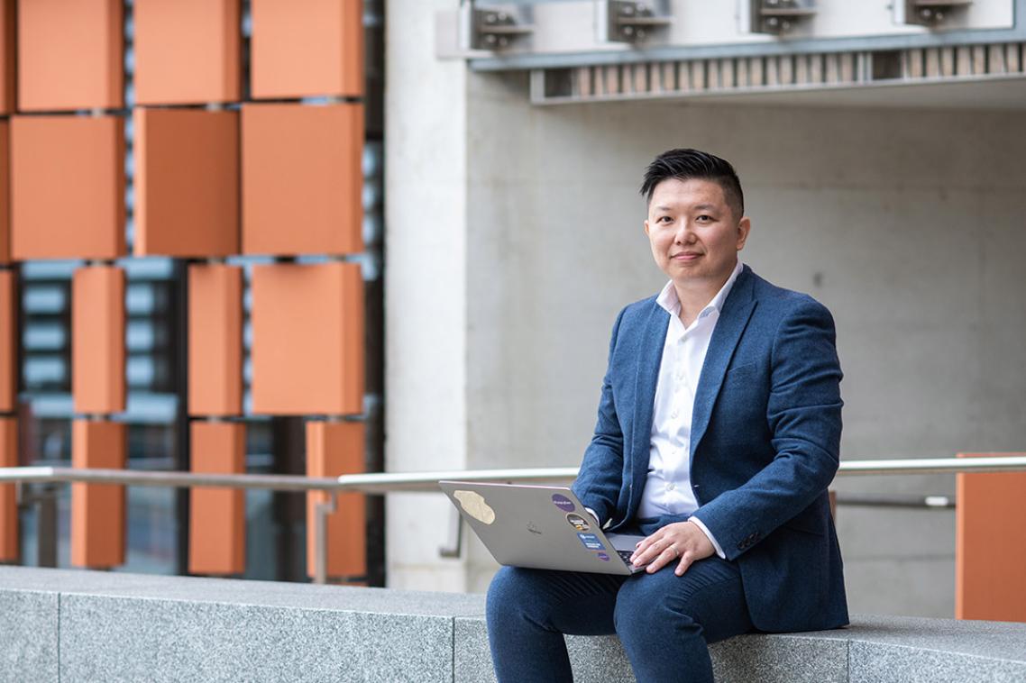 Professor Ryan Ko sitting with laptop in front of concrete building