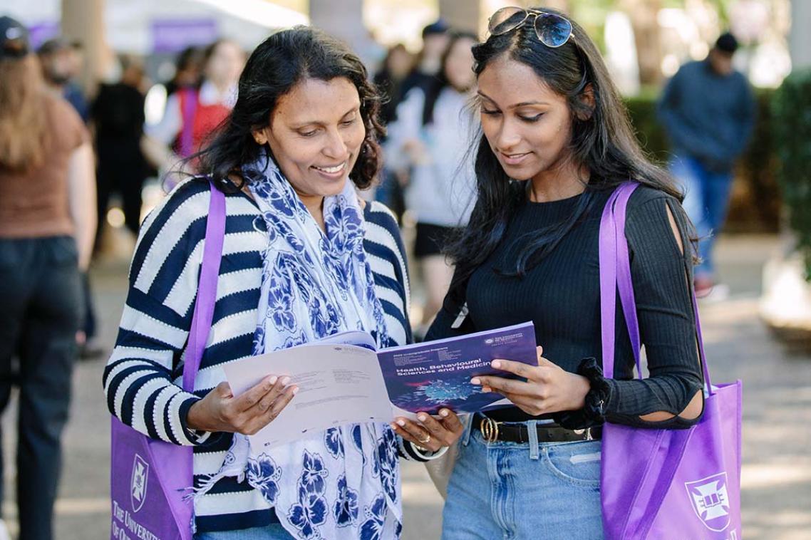 A mother and daughter at UQ Open Day looking at a booklet and holding purple UQ tote bags