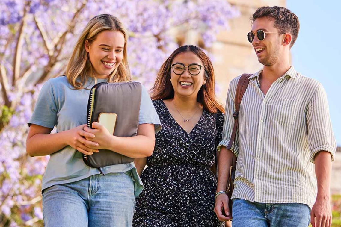 Three students walk through UQ St Lucia's Great Court smiling and chatting with jacaranda trees in the background