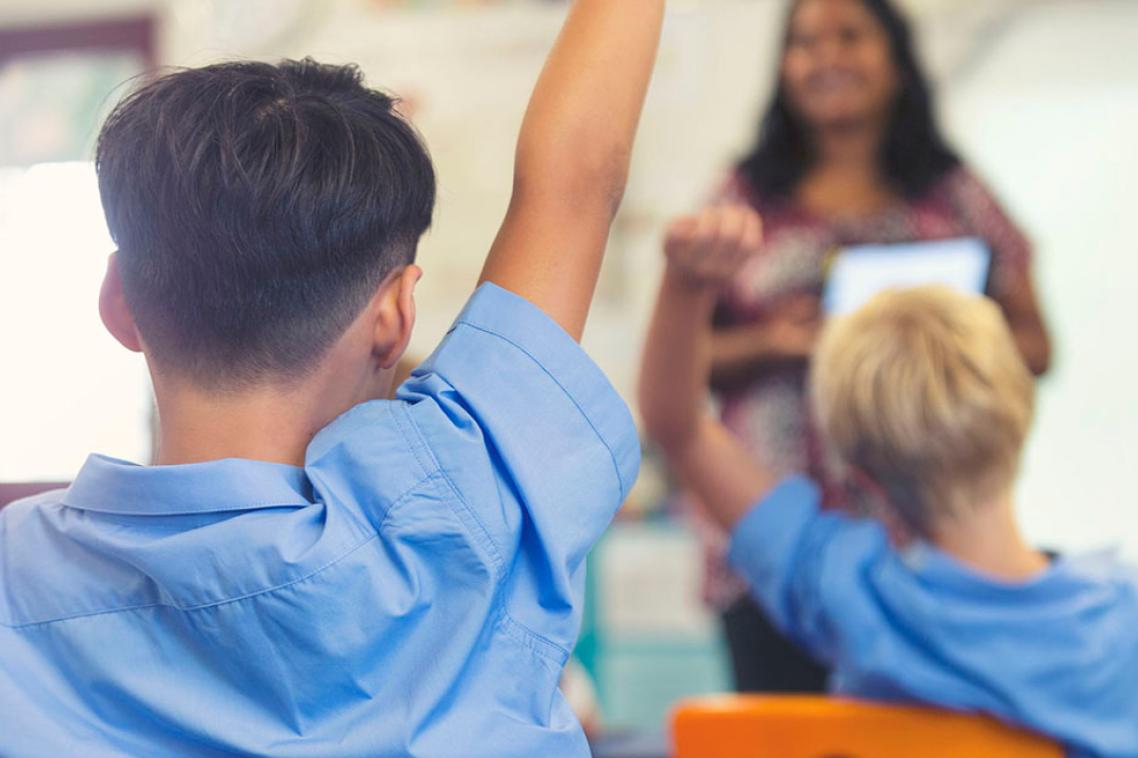 Primary school students raise their hand as a teacher stands in front of their classroom