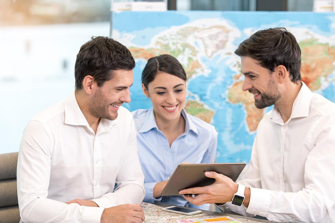 Three people in business clothes look at a tablet in front of a world map.