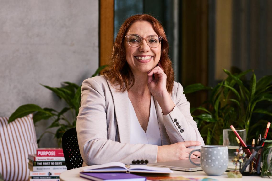 Kelly sits at a desk with multiple books in the background. 